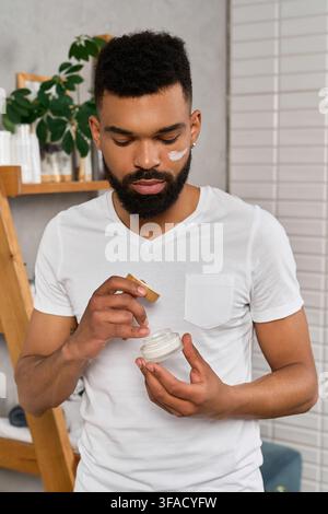 Ein junger Mann trägt vorsichtig Creme auf sein Gesicht auf, während er zu Hause steht. Stockfoto