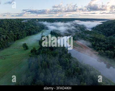Luftaufnahme des Gauja-Flusses, der sich durch Wald und grüne Wiesen schlängelt, die im Morgennebel bei Sigulda, Lettland, bedeckt sind. Stockfoto