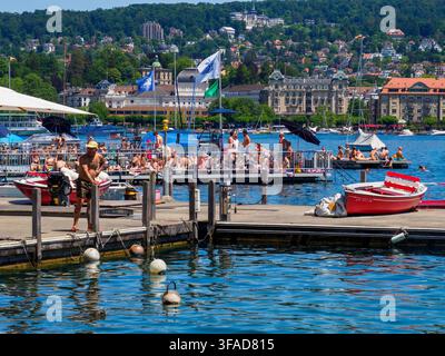 Breite panoramische Hochaufnahme der Zürcher Ufergegend, Bootverkehr und Schwimmer auf der Limmat sowie der historischen Skyline unter einem blauen Sommerhimmel. Stockfoto