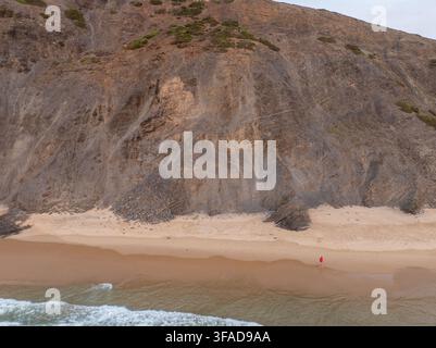 Luftaufnahme einer Person in einem roten Hemd, die entlang eines sandigen Strandes unter hoch aufragenden Felsen im Bezirk Faro, Portugal, läuft Stockfoto