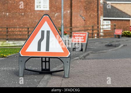 Ein dreieckiges Warnschild mit rotem Rand, das eine „Verengung der Straße“ anzeigt, steht im Vordergrund auf der Straße, mit Schildern „Gehweg gesperrt“ und „Fußweg gesperrt“ Stockfoto