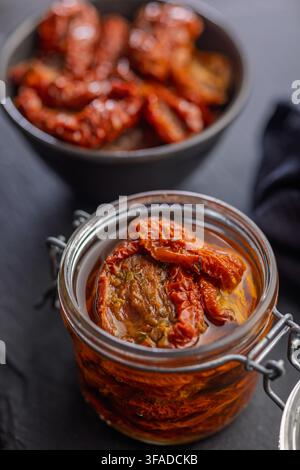 Glas mit sonnengetrockneten Tomaten in Olivenöl auf einer dunklen Schieferplatte, Nahaufnahme mit einer Schüssel Tomaten im Hintergrund; rustikal, geschmackvoll Stockfoto