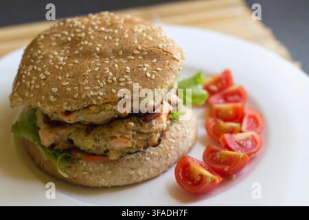 Hausgemachter Cajun-Lachsburger mit Salat auf einem Sesam-Vollkornbrötchen, halbierten Kirschtomaten auf einem weißen Teller, 45-Grad-Nahaufnahme Stockfoto