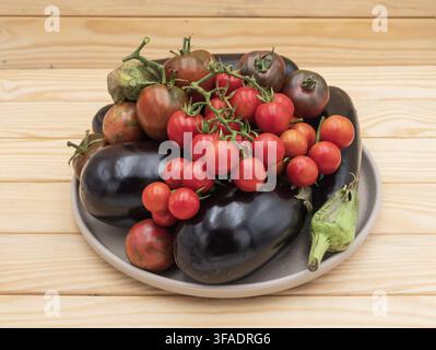 Glänzende violette Auberginen und verschiedene reife Tomaten sind auf einem Keramikteller gestapelt und bilden eine farbenfrohe Präsentation frischer saisonaler Gemüsesorten. Stockfoto