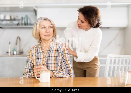 Eine alte Frau sitzt am Küchentisch und eine Frau mittleren Alters umschließt sie Stockfoto