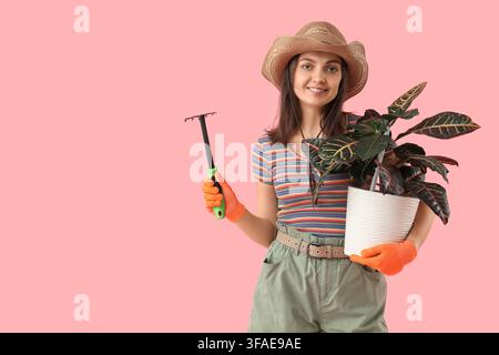 Gartenarbeit und Pflanzenpflege, junge Frau mit Strohhut, die eine Topfpflanze und einen Rechen auf einem rosa Hintergrund hält Stockfoto