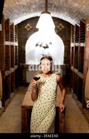 In einer charmanten Weinkeller hält eine Frau in einem stilvollen gepunkteten Kleid ein Glas Rotwein und strahlt während eines entzückenden Abends Raffinesse aus Stockfoto