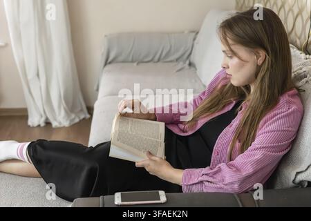Eine Frau sitzt auf einer Couch in einem Wohnzimmer. Sie liest ein Buch und ist entspannt. Ein Telefon liegt auf der Couch in der Nähe. Das Licht vom Fenster erhellt t Stockfoto