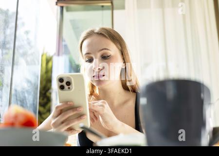 Eine junge kaukasische Frau mit langen blonden Haaren sitzt an einem Tisch und verwendet ein Smartphone. Eine Schale mit Obst und eine Tasse sind im Vordergrund sichtbar Stockfoto