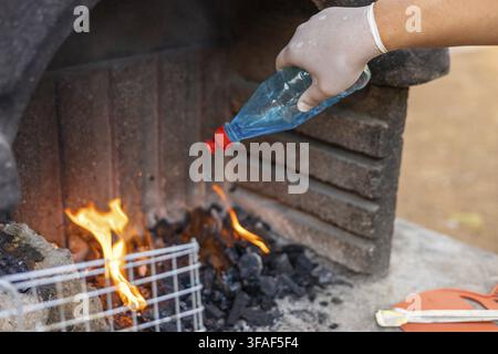 Eine behandschuhte Hand gießt blaue Flüssigkeit auf brennendes Holz in einem Stein-Grill. Flammen steigen aus der Glut auf, was die Vorbereitung zum Kochen anzeigt Stockfoto