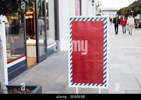 Ein leeres Mockup-Schild mit dekorativem Rand ist gut sichtbar vor einem Einzelhandelsgeschäft angebracht. Shopper schlendern vorbei, und die Sonne wirft Schatten in die Büste Stockfoto