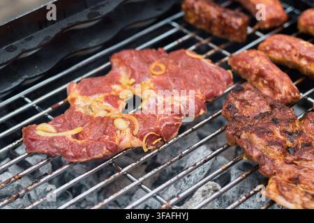 Nahaufnahme von gewürzten Schweinenackensteaks und rohen Würsten, die über brennenden Holzkohleglut auf einem Metallgrill gegrillt werden, um köstliche herzhafte Barbecue-Gerichte zuzubereiten Stockfoto