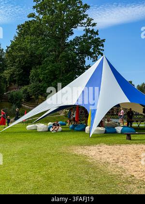 Riga, Lettland – 14. August 2026: Blauer und weißer Baldachin-Sonnenschutz auf einem sonnigen grünen Rasen aufgestellt, mit Leuten, die auf Sitzsäcken und Picknickbänken darunter entspannen Stockfoto