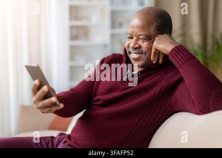 Lächelnder reifer schwarzer Mann entspannt sich auf dem Sofa, während er auf sein Smartphone schaut Stockfoto