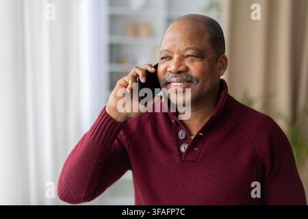 Lächelnder reifer schwarzer Mann, der zu Hause mit dem Smartphone telefoniert und das Gespräch genießt Stockfoto