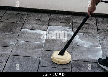 Mann reinigt schmutzige Patiofliesen mit einem Hochdruckreiniger im Freien, entfernt Moos und Schmutz von einem Wohnweg, Hauswartung, Außenreinigung Stockfoto