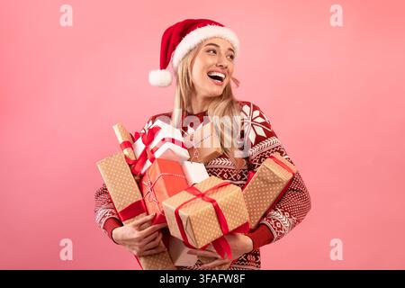 Schöne kaukasische Frau in Xmas Outfit halten Stapel von eingewickelten Weihnachtsgeschenke auf rosa Hintergrund. Urlaubsangebot Stockfoto