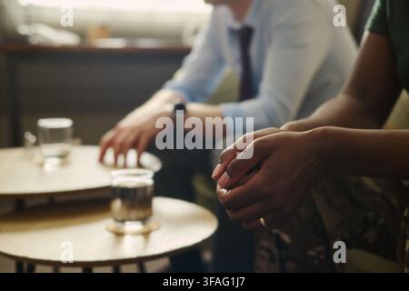 Afroamerikanische Frau sitzt mit kaukasischem Mann während einer Therapiesitzung, konzentriert auf verklammerte Hände. Bild repräsentiert psychologische Unterstützung und ist geeignet für Beratungsdienste Stockfoto