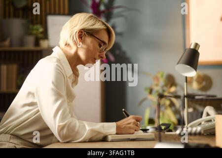 Mittelalte kaukasische Frau arbeitet am Bürotisch und macht Notizen in einem Notizbuch. Das Bild repräsentiert Unternehmensplanung und eignet sich für Business-Beratungs-Marketingmaterialien Stockfoto