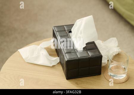 Schwarzer Leder-Taschentuchspender, der auf einem Holztisch neben einem Glas Wasser und zerknüllten Taschentüchern steht. Bild stellt die Erkältungs- und Grippesaison dar, geeignet für Gesundheitsmarketing Stockfoto