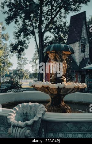 Verzierte Steinwasserspringbrunnen mit Puttenstatuen unter einem Regenschirm im öffentlichen Park Mogosoaia mit historischem Stilgebäude unscharf im Hintergrund Stockfoto