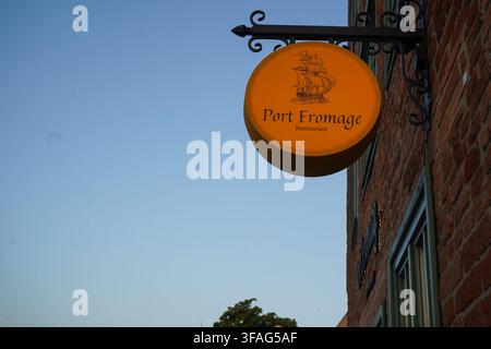 Niedrigwinklige Nahaufnahme eines beleuchteten gelben runden Schildes für das Port Fromage Restaurant, das an der Fassade eines historischen roten Backsteingebäudes bei Dämmerung angebracht ist. Stockfoto