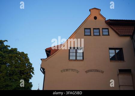 Niedrig angelegte Ansicht einer historischen Fassade aus beigem Putz mit Giebeldach und Gaubenfenstern neben einem grünen Baum in Danzig, Polen, vor klarem Himmel. Stockfoto
