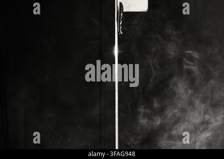 Abstrakter schwarzweißer Rauch, der sich sanft durch die Luft um ein dunkles strukturelles Element schlängelt, während Licht durch einen vertikalen Spalt fällt. Stockfoto