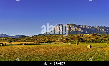 Landschaft mit felsigem Gebirgszug unter blauem Himmel. Felder mit goldenem Weizen erstrecken sich bis zu fernen Hügeln. Verstreute Heuballen ruhen auf einer sonnenbeschienenen Wiese. Gr Stockfoto