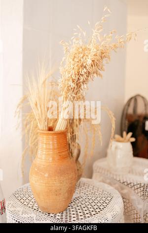 Eine Nahaufnahme eines Stilllebenarrangements mit getrocknetem Weizen in einer Terrakotta-Vase, platziert auf einem weißen Spitzentischtuch in einer gemütlichen Inneneinrichtung. Das Beige und Stockfoto