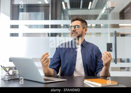 Bärtiger Mann mit Brille und blauem Hemd, der an einem modernen Bürotisch mit einem Laptop sitzt und aktiv an einer Diskussion oder Präsentation teilnimmt Stockfoto