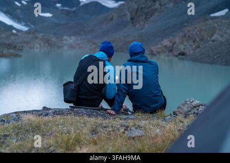 Zwei männliche Wanderer ruhen sich an einem Bergsee aus und prüfen die Karte auf dem Smartphone nach einer langen Wanderung in der Natur Stockfoto