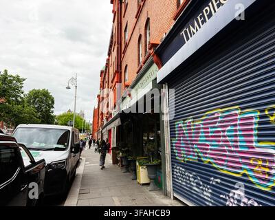 Dublin, Irland – 22. Mai 2026: Belebte Dubliner Straße mit graffitibedecktem Ladenschluss, geparkten Lieferwagen, Backsteingebäuden und Fußgängern, die entlanggehen Stockfoto
