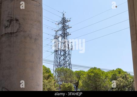 Ein großer Metall-Hochspannungsmast steht hoch und überträgt Strom über zahlreiche Drähte über einer Reihe üppiger grüner Bäume hinweg Stockfoto