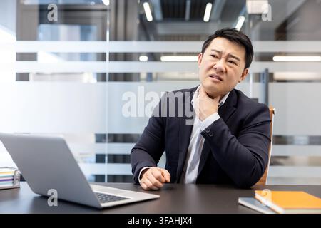 Asiatischer Geschäftsmann sitzt am Schreibtisch im Büro, erlebt starke Halsschmerzensschmerzen, hält seinen Hals und drückt Unbehagen aus, während er an einem Laptop arbeitet Stockfoto