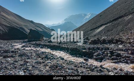 Ein Bach fließt durch ein felsiges Tal in Kirgisistan. Hohe Berge erheben sich im Hintergrund. Es ist Mittag mit hellem Sonnenlicht, das die Landschaft erhellt Stockfoto