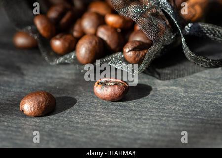 Geröstete Kaffeebohnen quellen aus einem Netzbeutel auf eine dunkle strukturierte Oberfläche. Stockfoto