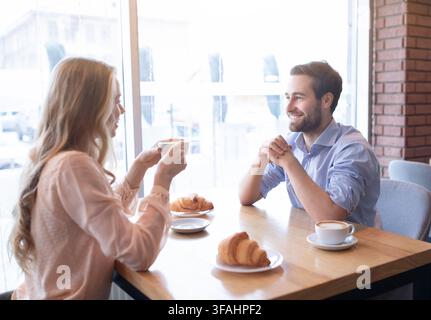 Liebendes junges Paar, das in der Nähe des Fensters in einem Café zu Mittag isst, aromatischen Kaffee trinkt und sich unterhält Stockfoto