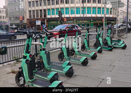 Eine Reihe grüner Bolt-Elektro-Scooter, die auf einem Gehweg in Liverpool, UK, geparkt sind, mit Stadtgebäuden, einem roten Auto und einer Ampel im Hintergrund. Stockfoto