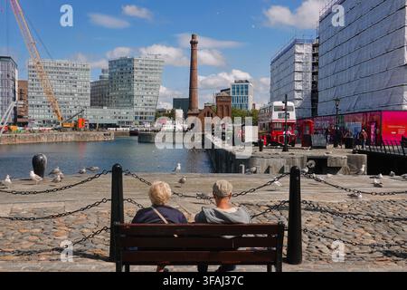 Rückansicht eines Seniorenehepaars, das auf einer Bank am Royal Albert Dock in Liverpool, UK, sitzt und über das Wasser zum The Pump House Pub schaut Stockfoto
