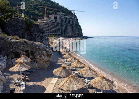 Schattige leere Plastik-Sonnenliegen unter Strohschirmen auf einem Kiesstrand mit Blick auf eine malerische Landschaft aus felsigen Klippen und Mittelmeer Stockfoto