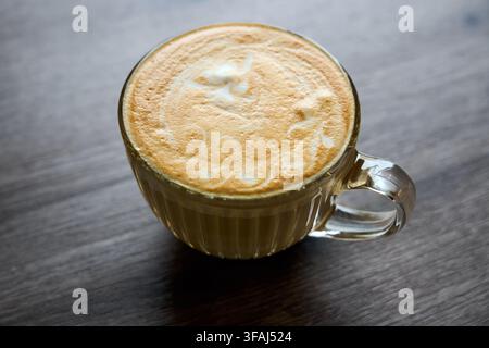 Frisch gebrühter Cappuccino mit detaillierter Schaum-Latte-Art, heiß in einem klaren gerippten Glasbecher serviert, auf dunklem, texturiertem Holztablett-Hintergrund, perfekt für Brea Stockfoto