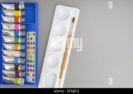 Wasserfarbtuben in einem blauen Organizer mit einer weißen Palettenablage und einem kleinen Pinsel, der auf einem flachen grauen Hintergrund mit negativem Raum für Text liegt. Stockfoto