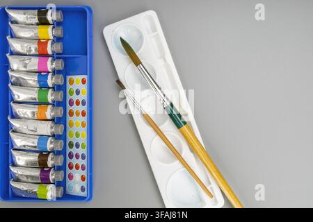 Oberansicht von bunten Aquarellfarbtuben, zwei Holzpinsel und einer weißen Mischpalette, ordentlich angeordnet auf einer sauberen, minimalen grauen Tischfläche. Stockfoto