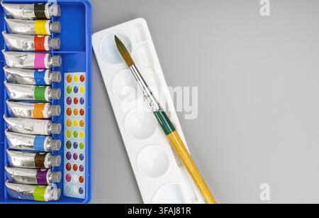 Flache Ansicht von bunten Aquarellfarbtuben, weißer Plastikmalpalette und Holzpinsel, die auf einem glatten grauen Hintergrund mit Kopierplatz liegen. Stockfoto