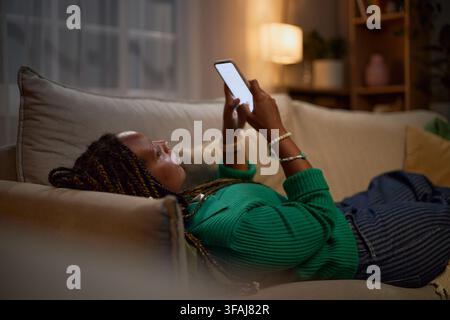 Schwarze Frau liegt auf dem Sofa und benutzt nachts ein Smartphone Stockfoto