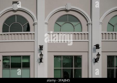 Symmetrische Frontansicht einer weißen Gebäudefassade mit dekorativen Bogenfenstern, Gitterglasfenstern, Vintage-Wandlampen und weißem Architekturpfeiler Stockfoto
