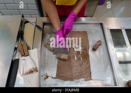 Hände in hellrosa Gummihandschuhen führen ein Metallspachtelwerkzeug, um kalte Schokoladen-Milchmischung auf gefrorener Stahlarbeitsplatte mit Holzschabern und Papier zu rollen Stockfoto