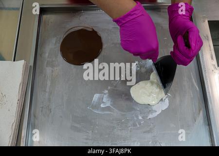 Hände in hellrosa Gummihandschuhen verwenden Stahlspatel, um braune Kakaopaste und weiße Milchcremeberge auf einer gefrorenen Edelstahl-Arbeitsfläche zu vermischen, Ansicht von oben Stockfoto