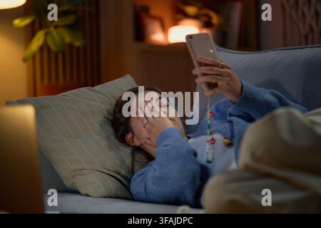 Müde Frau liegt auf dem Sofa und verwendet nachts ihr Smartphone Stockfoto